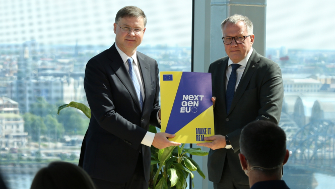 Divi vīrieši tumšos uzvalkos stāv plecu pie pleca, rokās turot liela formāta mapi, fonā pilsētas skats