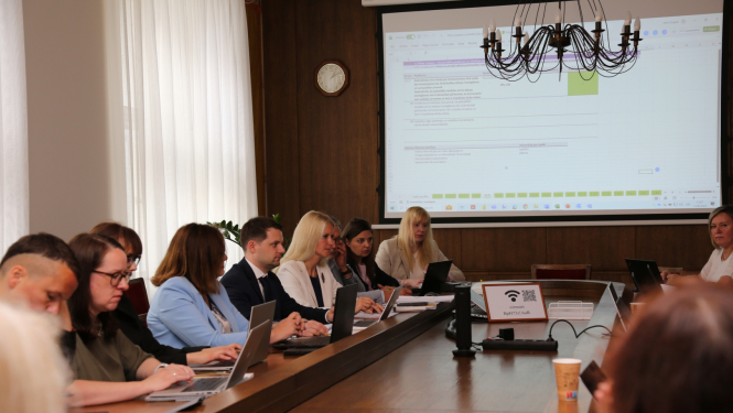 pie sanāksmju galda sēž vīrieši un sievietes un notiek savstarpējas diskusijas