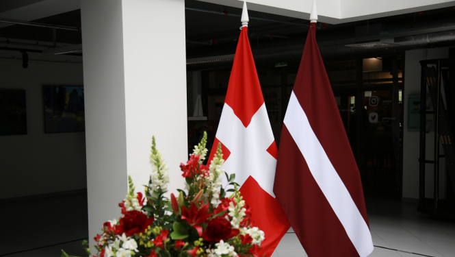 Latvijas un Šveices karogi uz statīviem, priekšplānā ziedu pušķis