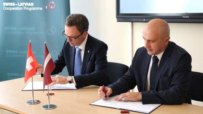 Divi vīrieši sēž pie galda un paraksta dokumentus, uz galda priekšplānā stāv Latvijas un Šveices karodziņi