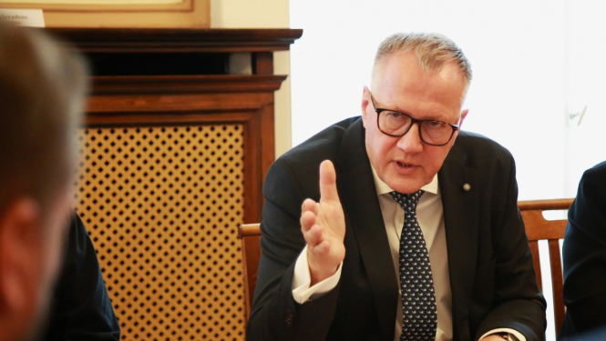 Vīrietis uzvalkā ar paceltu roku sarunājas ar cilvēku otrpus galdam