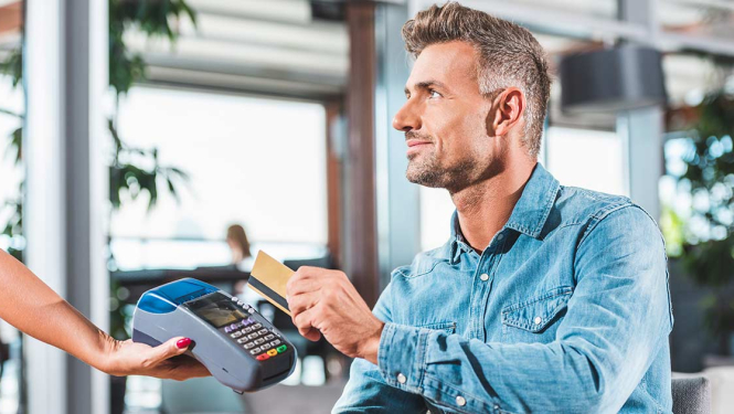 Vīrietis zilā kreklā āra terasē nomaksā rēķinu par ēdināšanas pakalpojumu