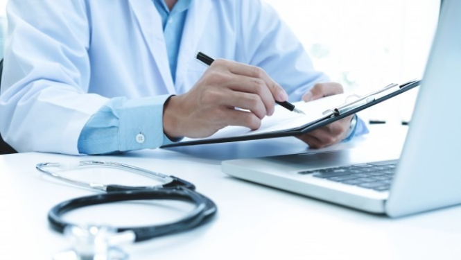 Ārsts pie galda ar datoru un pierakstu mapi, pildspalvu