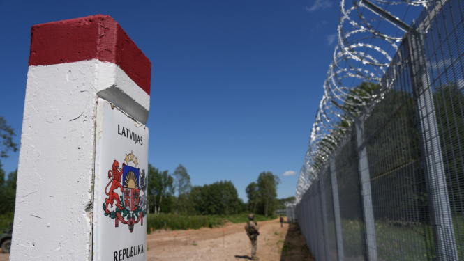 Attēlā tuvplānā redzams robežstabiņš ar Latvijas ģerboni, līdzās augstam žogam ar dzeloņstieplēm