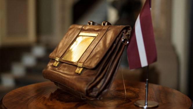 Attēlā redzams tradicionālais budžeta portfelis statīvā novietots uz apaļa galdiņa, līdzās stāv Latvijas karodziņš