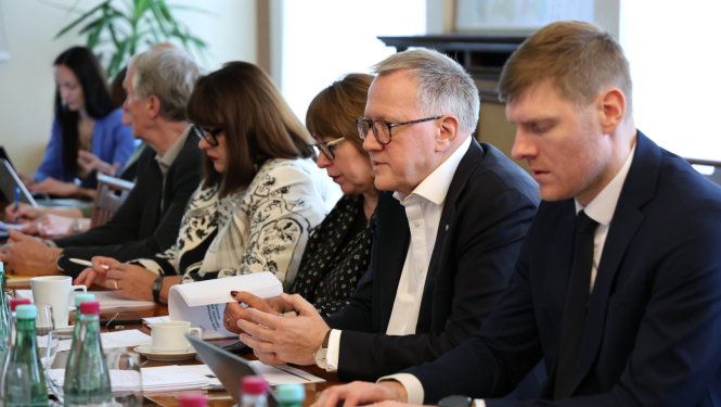 Formāli ģērbušos cilvēku grupa sēž pie galda un piedalās sanāksmē. Daži strādā ar datoru, citi pēta dokumentus. Uz galda stāv glāzes un ūdens pudeles