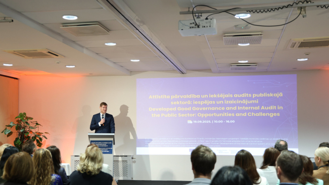 Attēlā auditorijas priekšā pie tribīnes stāv vīrietis uzvalkā ar mikrofonu rokās, uz sienas ir ekrāns, uz kura redzama prezentācija