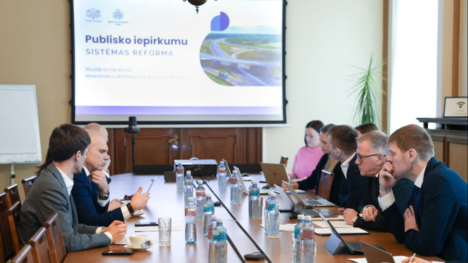 Pie gara sēžu galda abās pusēs sēž vīrieši un sievietes, savstarpēji diskutē, attēla centrā projektora ekrāns, uz kura redzama prezentācija 