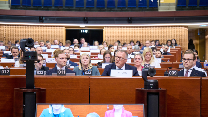 Attēlā redzams plenārsēžu zāles iekšskats ar dalībniekiem, kas sēž pie galdiem un uzklausa runātāju. 