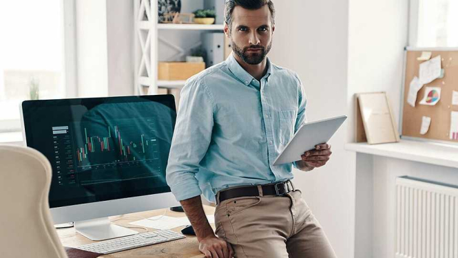 Attēlā redzams vīrietis ar planšetdatoru rokā, atspiedies pret darbagaldu.