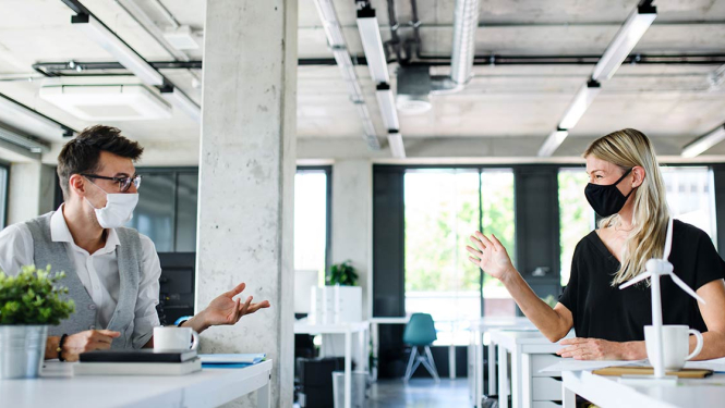 Attēlā divi kolēģi sarunājas, uzvilkuši sejas aizsargmaskas