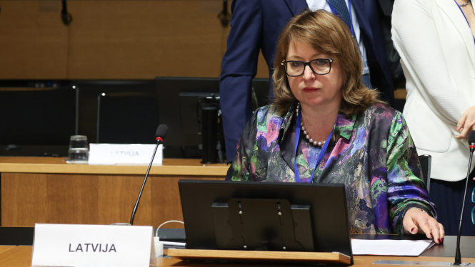 Sieviete krāsainā blūzē sēž pie sanāksmju galda, uz tā novietots dators, dokumenti un mikrofons