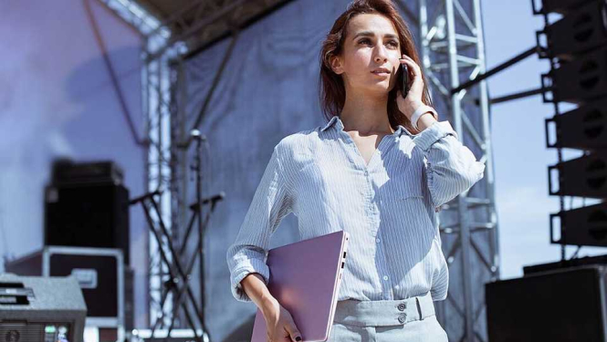 Attēlā redzama sieviete, kura runā pa telefonu, turot datoru otrā rokā