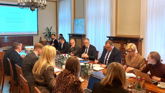 Zālē pie gara galda sēž vīrieši un sievietes un savstarpēji sarunājas