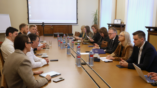 Pie garena galda sēž divas cilvēku grupas, kas apspriež darba jautājumus. Uz galda redzami klēpjdatori, dokumenti, telefoni un ūdens pudeles, fonā projekcijas ekrāns