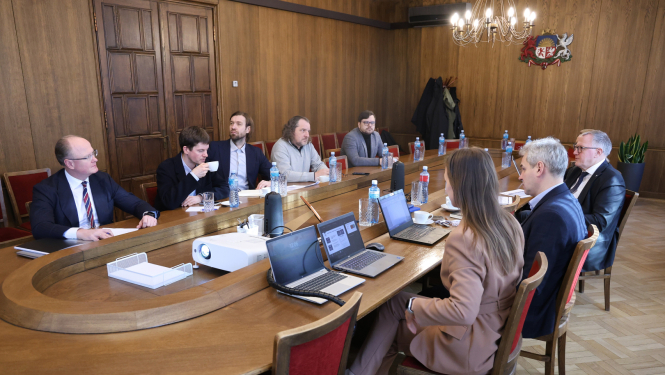 Pie sēžu galda sēž vīrieši un sieviete, savstarpēji diskutē, uz galda stāv dokumenti un portatīvie datori