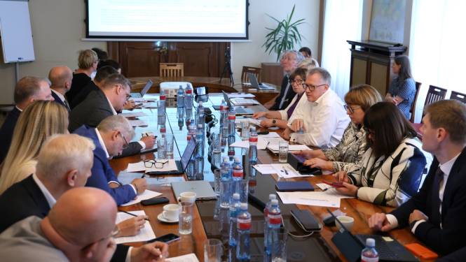 Attēlā redzama sanāksme, kurā ap lielu galdu sēž vairāki cilvēki, uz galda novietoti dokumentiem un klēpjdatoriem, pie sienas redzams liels ekrāns ar prezentāciju