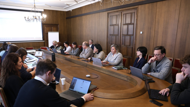 Pie garena sanāksmju galda sēž sievietes un vīrieši, un diskutē, uz galda stāv portatīvie datori un dokumenti