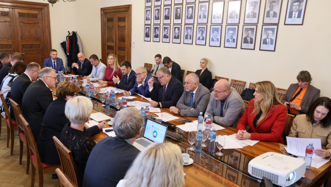 Attēlā garš galds ap kuru no visām pusēm sēž vīrieši un sievietes un savstarpēji sarunājas, uz galda stāv dokumenti, portatīvie datori, ūdens pudeles un glāzes