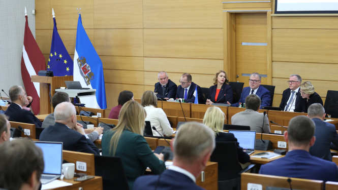 Pie gara galda auditorijas priekšā sēž seši cilvēki, kuri diskutē ar auditoriju, telpas malā stāv Latvijas ES un Rīga domes karogi
