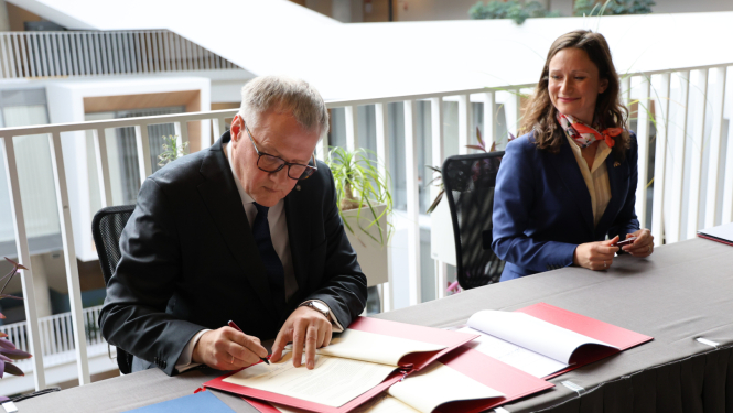 Attēlā vīrietis paraksta uz galda stāvošus dokumentus, blakus sēdošā sieviete skatās uz viņu