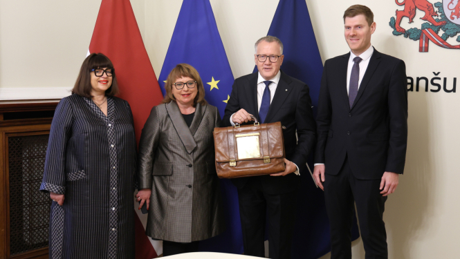 Attēlā uz Latvijas, ES un Nato karogu fona stāv četri cilvēki un smaida, divi no tiem rokās tur portfeli