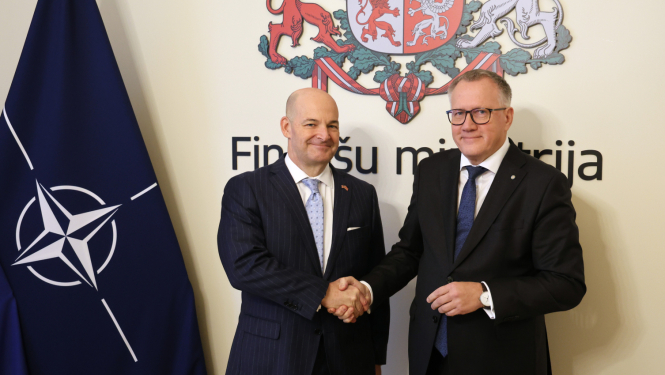 Attēlā uz Finanšu ministrijas ģerboņa fona, sarokojas divi vīrieši uzvalkos, blakus redzams NATO karogs