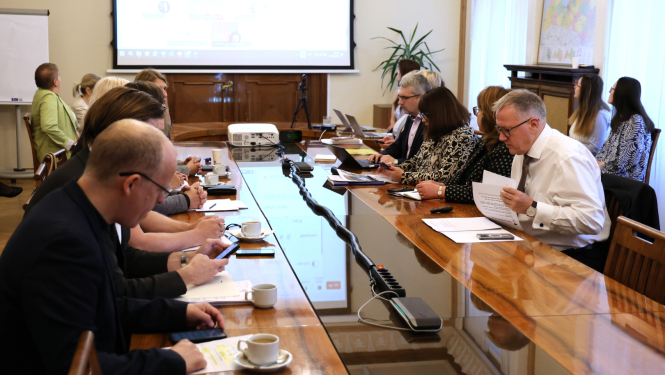 Sanāksmes laikā cilvēki sēž pie gara koka galda, skatās dokumentus, lieto datorus; fonā redzams projekcijas ekrāns ar prezentāciju