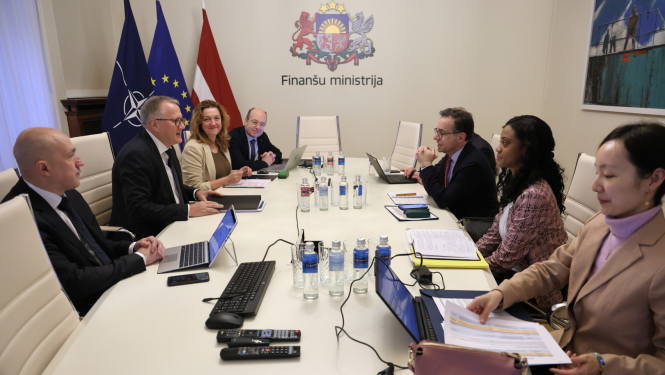 Attēlā redzama sanāksmju telpa Finanšu ministrijā: ap galdu sēž ministrijas pārstāvji un delegācijas locekļi, uz galda redzami klēpjdatori un dokumenti, fonā redzami NATO, ES un Latvijas karogi un uz sienas uzraksts “Finanšu ministrija”