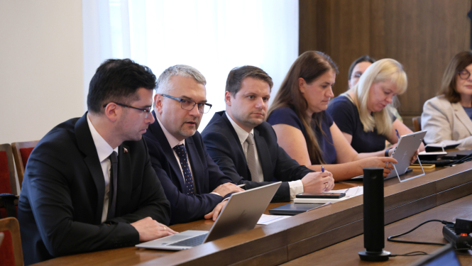 Pie sēžu galda rindā sēž vīrieši un sievietes, savstarpēji sarunājas. Uz galda novietoti portatīvie datori un dokumenti.