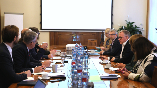 Pie gara galda no abām pusēm sēž vīrieši un sievietes, savstarpēji sarunājas, pie sienas redzams ekrāns, uz kura rāda prezentāciju