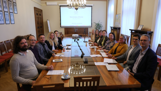 Attēlā pie gara galda abpus galdam sēž vīrieši un sievietes un smaidot skatās kamerā, tālumā pie sienas ir redzams projektora ekrāns