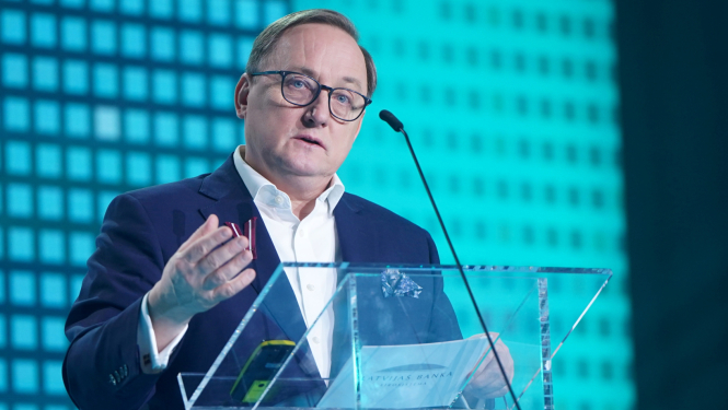 Attēlā tuvplānā redzams vīrietis, kurš stāv pie caurspīdīgas tribīnes un žestikulējot runā, viņas priekšā ir mikrofons