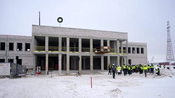 Attēlā redzama daļēji uzcelta divstāvu sabiedriskā ēka ziemas apstākļos; priekšplānā sniegotā teritorijā stāv cilvēku grupa dzeltenās drošības vestēs un ķiverēs, notiek būvlaukuma apskate