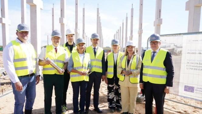 Bildē redzama cilvēku grupa būvlaukumā, tērpušies dzeltenās atstarojošās vestēs un aizsargķiverēs, fonā redzami būvkonstrukciju balsti un smilšaina zeme. Vienam no cilvēkiem rokās ir kapsula, kas paredzēta pamatakmens iemūrēšanas ceremonijai.