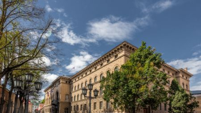 Saeimas ēka, priekšplānā zaļo koki, zilas, mākoņainas debesis