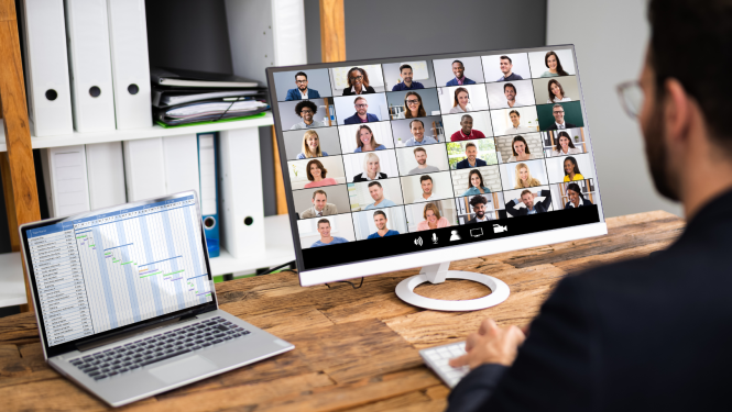 Vīrietis redzams no mugurpuses, sēž iepretim datora ekrānam, kurā redzamas cilvēku sejas