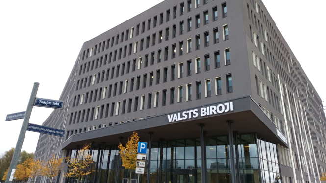 Attēlā redzama pelēka daudzstāvu biroju ēka ar stiklotu ieejas fasādi, uz tās redzams uzraksts VALSTS BIROJI