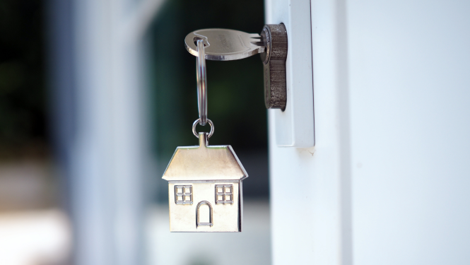 Attēlā redzama atslēga, kas ievietota slēdzene, pie tās karājas metāla atslēgu piekariņš mājas veidolā