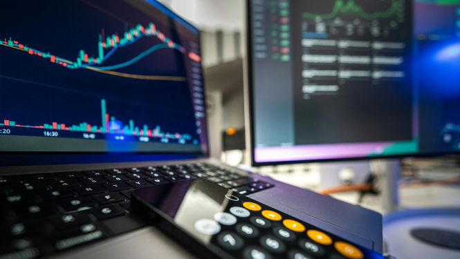 Attēlā redzams datora ekrāni, kuros redzamas akciju tirgus diagrammas ar cenu svārstībām, priekšplānā klēpjdators, kalkulators un tastatūra