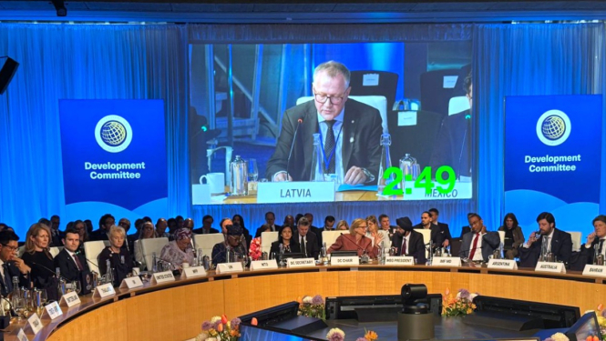 Konferenču zālē pie apaļa galda sēž delegāti no dažādām valstīm, ekrānā redzams finanšu ministrs uzstājoties ar runu