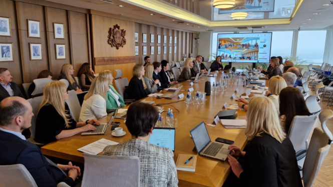 Pie gara galda sēž vīrieši un sievietes, savstarpēji sarunājas, uz galda stāv dokumenti un portatīvie datori, telpas galā ir redzams ekrāns uz kura tiek rādīta prezentācija