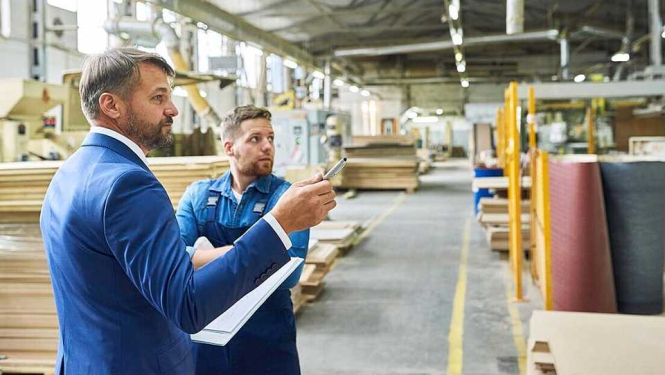 Attēlā divi vīrieši rūpnīcas telpās sarunājas