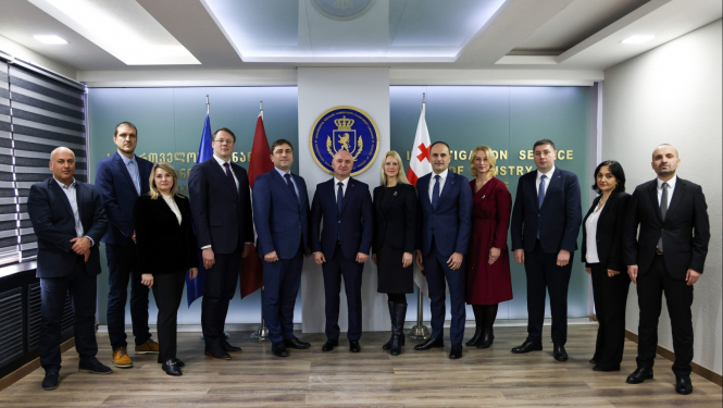 Uz ģērboņa un karogu fona rindā sastājušies stāv vīrieši un sievietes 