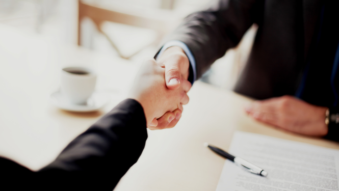 Virs galda virsmas redzamas cilvēku rokas, kuras sarokojas, uz galda stāv dokumenti un kafijas tasīte