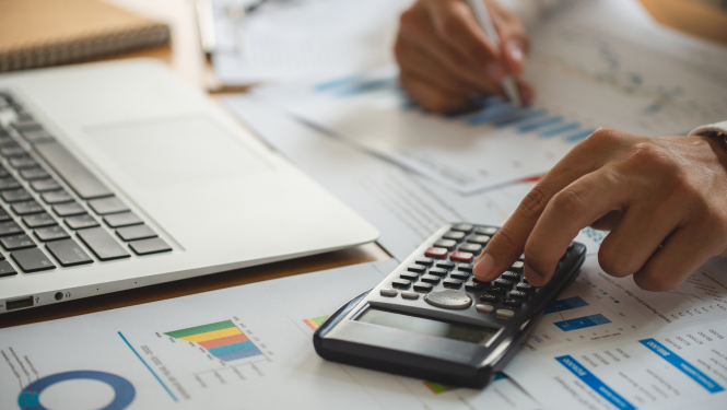 Tuvplānā galds ar dokumentiem, redzamas cilvēka rokas, kas rēķina izmantojot kalkulatoru un veicot pierakstus