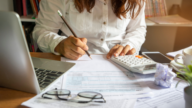 sieviete baltā blūzē sēž pie galda un ar zīmuli veic aprēķinus. uz galda redzami dators, dokumenti un kalkulators