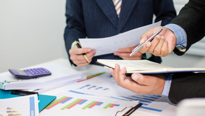 Pie galda sēž divi vīrieši un caurskata dažāda veida dokumentus