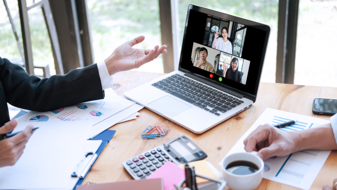 Uz darba galda stāv atvērts portatīvais dators un dažādi dokumenti un biroja elementi, redzamas cilvēku, kuri sēž pie galda, rokas