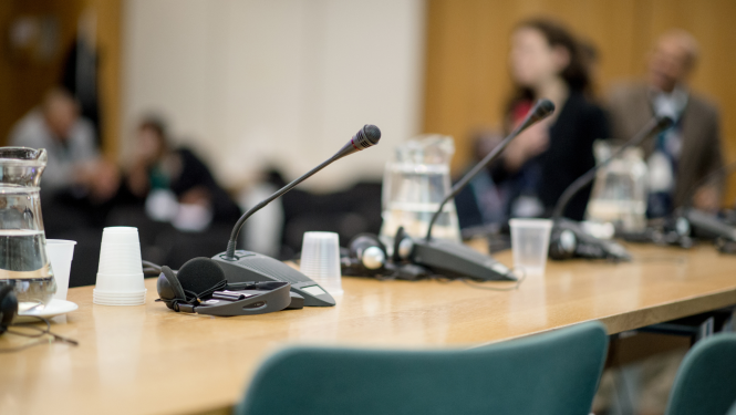 Tuvplānā redzama galda virsma uz kuras stāv galda mikrofoni un ūdens karafe ar glāzē, tālumā redzami cilvēku situeti